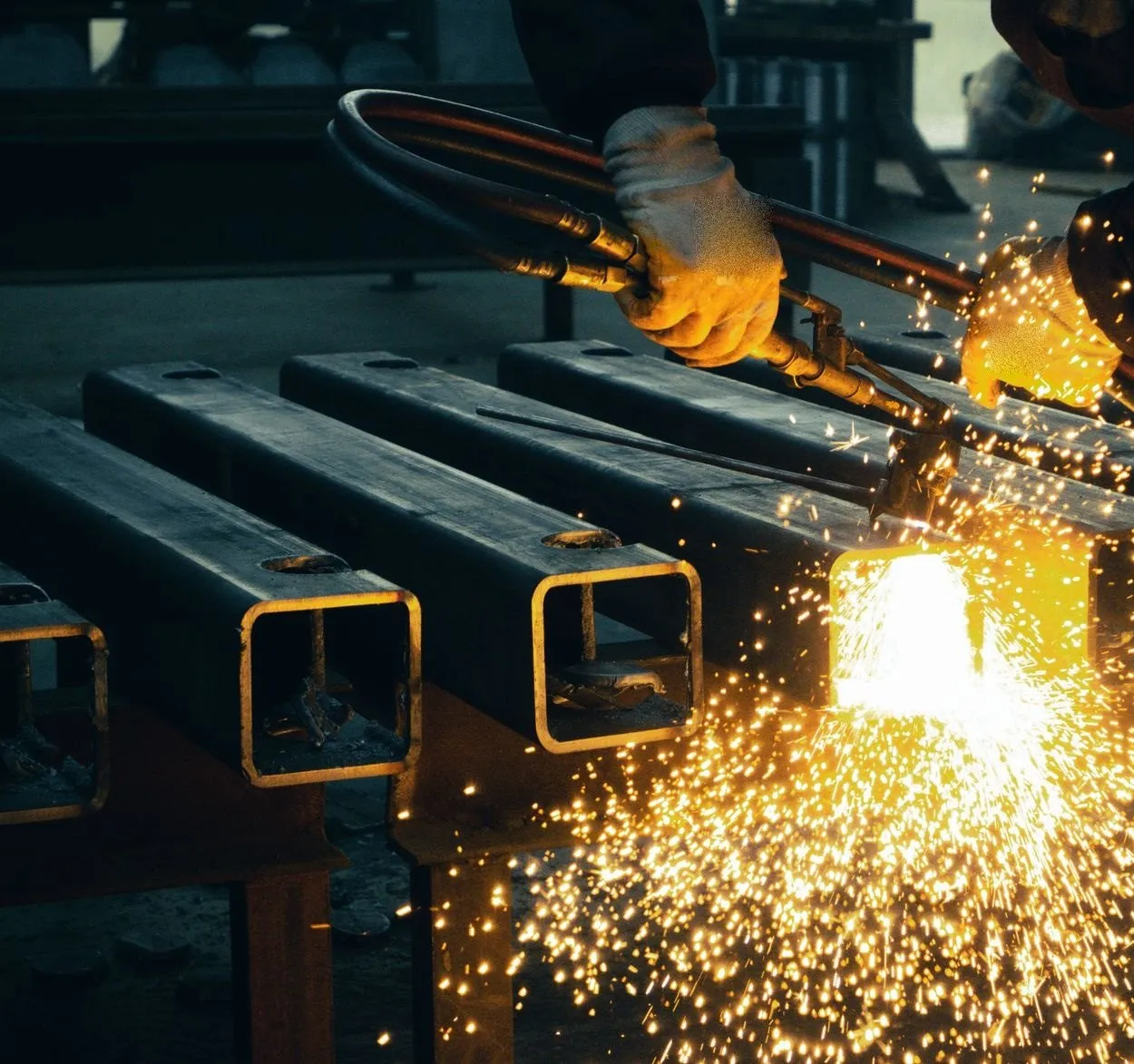 Gloved hands cutting box section with a torch, sparks spraying across the bench
