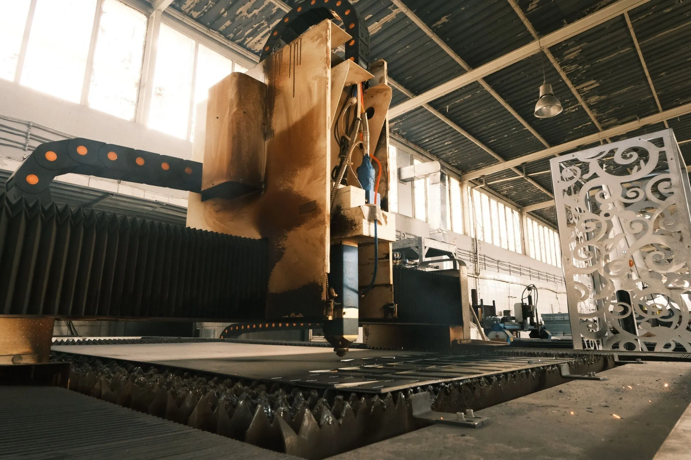 The fibre laser gantry over a part-cut steel sheet in the daylit cutting bay, a laser-cut decorative screen standing beside it