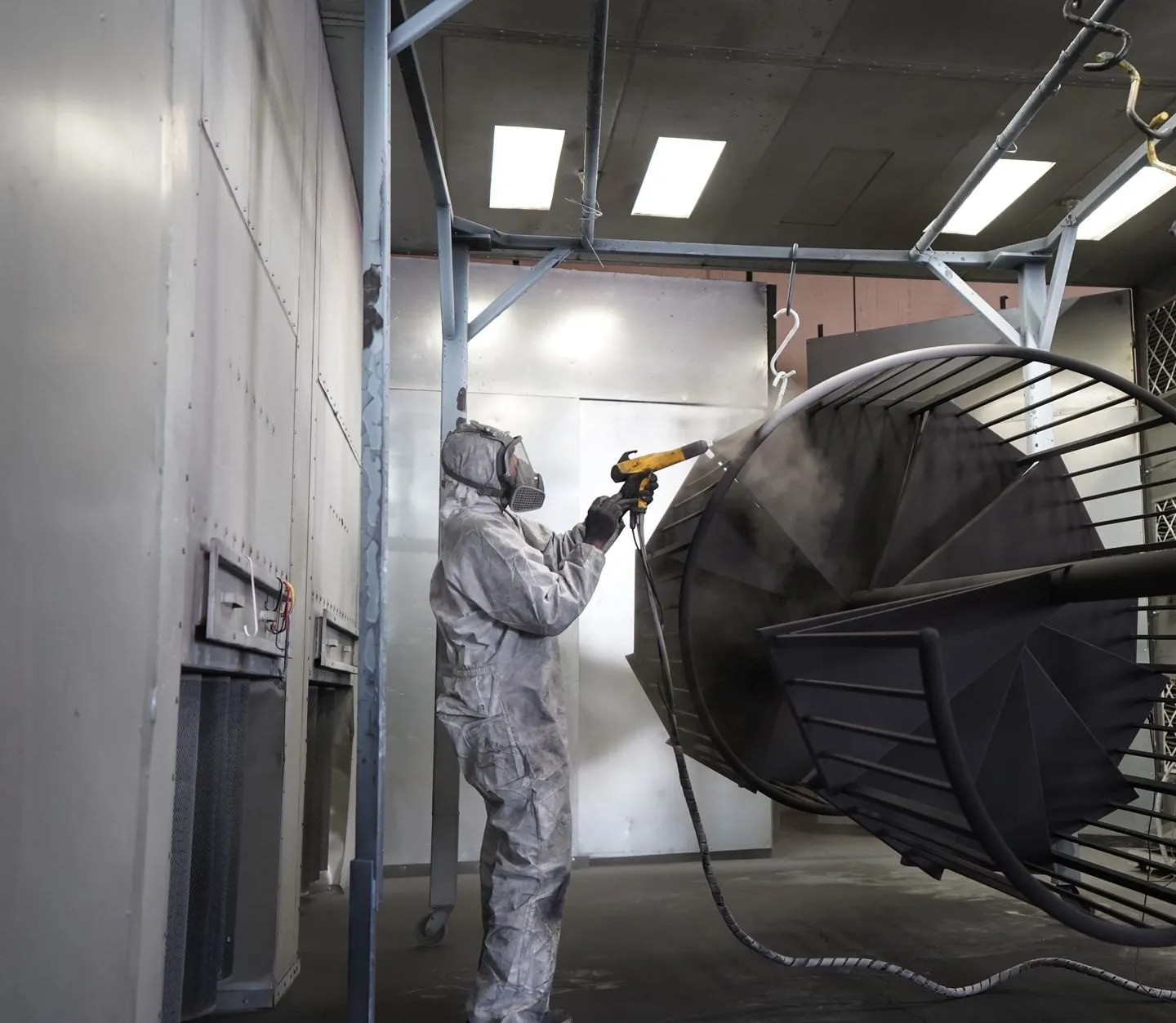 A painter in a protective suit and respirator spraying powder onto a spiral stair hung in the booth