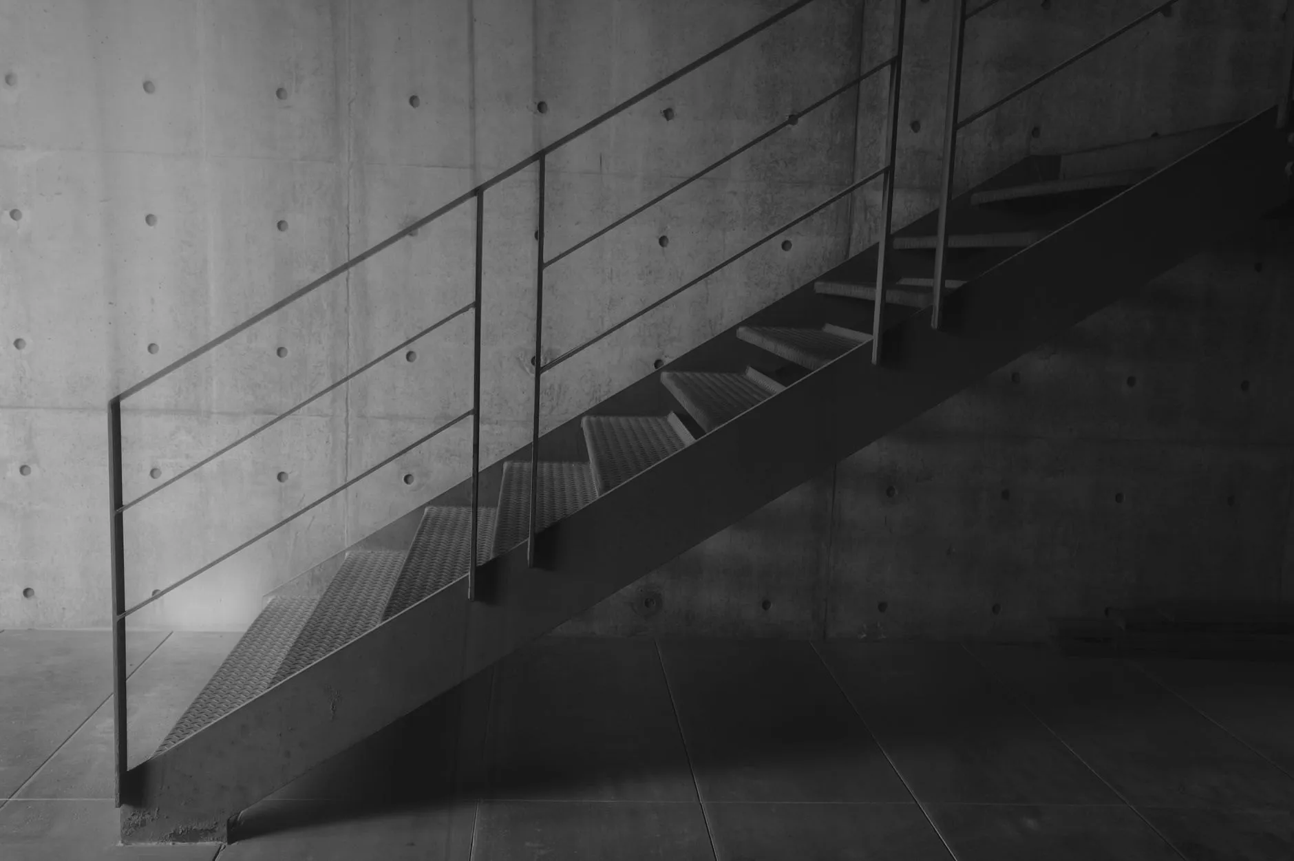A slim black steel stair with a bar handrail against a concrete wall