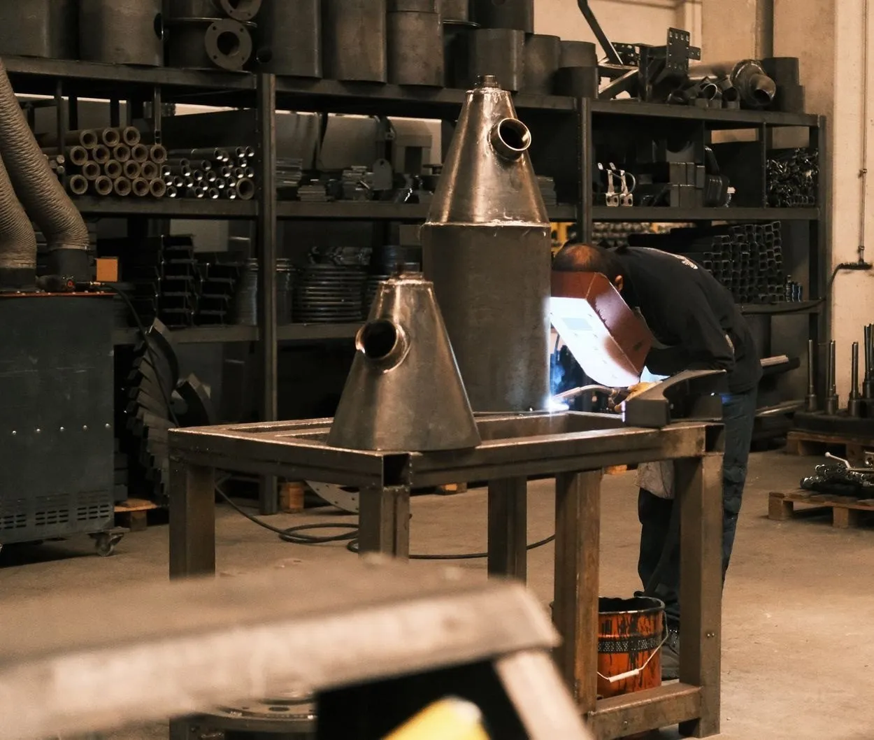 A welder in a helmet working at a steel bench beside two fabricated cone sections